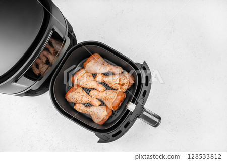 Raw seasoned chicken drumsticks are neatly arranged in the basket of a black air fryer, ready to be cooked on a clean white kitchen counter 128533812