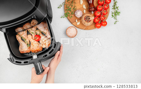 Raw marinated chicken drumsticks with herbs and a cherry tomato are arranged in an air fryer basket, surrounded by fresh ingredients like vine tomatoes, garlic, salt, and pepper on a kitchen counter 128533813
