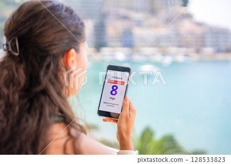 Young woman checking UV index on smartphone with high radiation alert. Mobile app warns to apply sunscreen before going outdoors 128533823