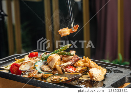 Chopsticks lift a steaming shrimp from a sizzling seafood platter featuring mussels, scallops, vegetables, and creamy sauce on a hot grill plate 128533881