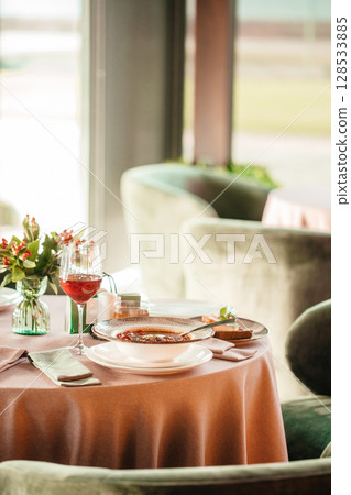 Elegant restaurant table setting with a bowl of hot soup, wine glass, and floral decoration in warm natural light 128533885