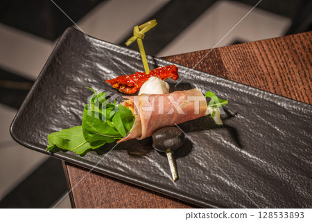 Elegant appetizer with prosciutto-wrapped arugula, mozzarella, sundried tomato, and black olive on a black ceramic plate 128533893