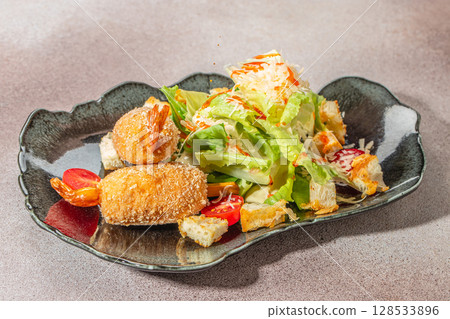 Crispy breaded shrimp served with fresh Caesar salad, cherry tomatoes, croutons, grated cheese, and dressing on a black plate Crispy breaded shrimp served with fresh Caesar salad, cherry tomatoes, croutons, grated cheese, and dressing on a black plate 128533896