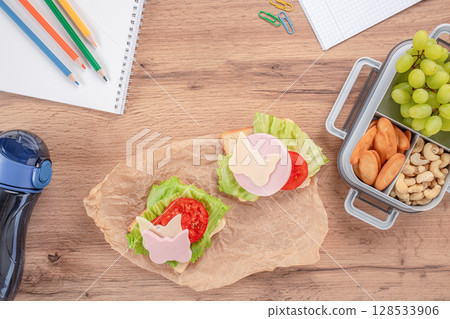Creative school lunch with open sandwiches decorated with butterfly-shaped cheese and ham, served alongside grapes, nuts, and crackers in a lunchbox on a wooden desk Creative school lunch with open sandwiches decorated with butterfly-shaped cheese and ham, served alongside grapes, nuts, and crackers in a lunchbox on a wooden desk 128533906