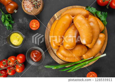 Round wooden plate filled with smoked sausages surrounded by fresh tomatoes, herbs, sauces, and spices on a dark stone background Round wooden plate filled with smoked sausages surrounded by fresh tomatoes, herbs, sauces, and spices on a dark stone background 128533911