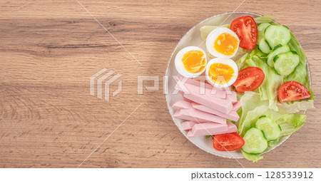 Healthy balanced plate with soft-boiled eggs, ham strips, fresh lettuce, tomatoes, and cucumber slices on a wooden table background. top view. copy space 128533912