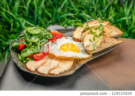 Nutritious breakfast plate with a sunny-side-up egg, fresh spinach and tomato salad, sliced chicken breast, and whole grain toast topped with hummus and herbs 128533920
