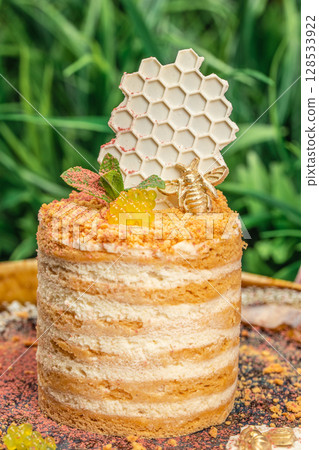 Elegant honey cake with layered cream and sponge, topped with white chocolate honeycomb, golden bee, mint leaves, and decorative spheres, set against a natural green background 128533922