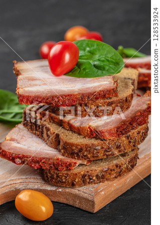 Stacked rye bread slices topped with spicy pork belly, fresh spinach leaves, and cherry tomatoes, served on a rustic wooden board Stacked rye bread slices topped with spicy pork belly, fresh spinach leaves, and cherry tomatoes, served on a rustic wooden board 128533924