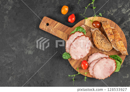 Sliced smoked pork is beautifully arranged with fresh spinach, multigrain bread, and colorful cherry tomatoes on a rustic wooden serving board 128533927