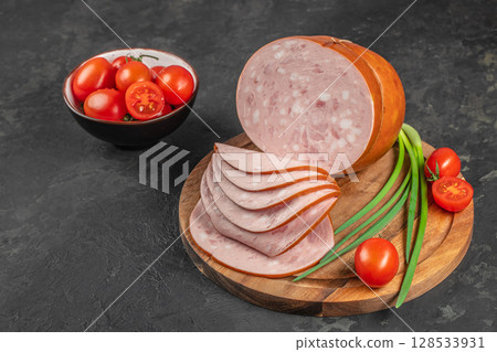 Slices of traditional bologna sausage are artfully arranged on a wooden board beside fresh cherry tomatoes and green onions on a dark stone background 128533931