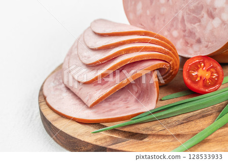Thin slices of classic bologna sausage with a smooth texture and orange rind rest neatly on a wooden cutting board, accompanied by fresh green onions and a halved cherry tomato 128533933