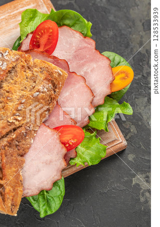 A rustic sandwich made with hearty whole-grain bread, fresh lettuce, tomato slices, and savory smoked ham creates a wholesome and appetizing meal on a dark stone background 128533939
