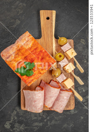 Sliced ham with a paprika crust is elegantly arranged on a wooden board alongside cheese cubes, green olives, and cocktail skewers, creating a vibrant and appetizing charcuterie presentation Sliced ham with a paprika crust is elegantly arranged on a wooden board alongside cheese cubes, green olives, and cocktail skewers, creating a vibrant and appetizing charcuterie presentation 128533941