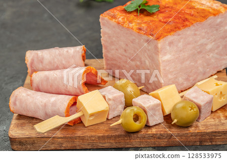 Rolled ham slices, cheese cubes, olives, and skewered appetizers are arranged on a wooden board next to a large block of ham loaf for a hearty and inviting snack display Rolled ham slices, cheese cubes, olives, and skewered appetizers are arranged on a wooden board next to a large block of ham loaf for a hearty and inviting snack display 128533975