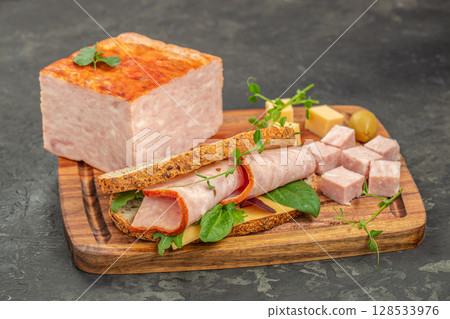 A hearty sandwich with rolled ham slices, cheese, and greens is served on a wooden board next to a block of ham loaf and diced ham cubes for a rustic and satisfying meal 128533976