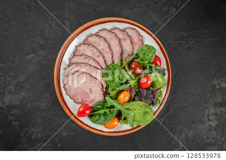 Thin slices of seasoned ham are arranged on a plate with a colorful fresh salad of leafy greens and cherry tomatoes for a light and appetizing dish 128533978