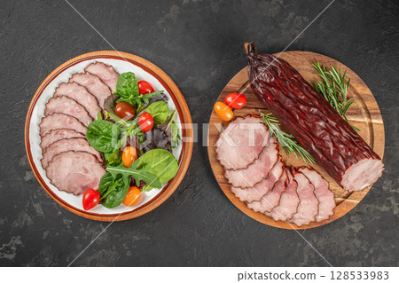 Two beautifully arranged plates display sliced cured meat with vibrant leafy greens, cherry tomatoes, and fresh rosemary on a dark rustic background Two beautifully arranged plates display sliced cured meat with vibrant leafy greens, cherry tomatoes, and fresh rosemary on a dark rustic background 128533983