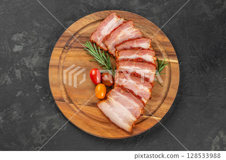Slices of spicy smoked pork belly arranged in a fan shape on a wooden board with fresh rosemary and colorful cherry tomatoes Slices of spicy smoked pork belly arranged in a fan shape on a wooden board with fresh rosemary and colorful cherry tomatoes 128533988