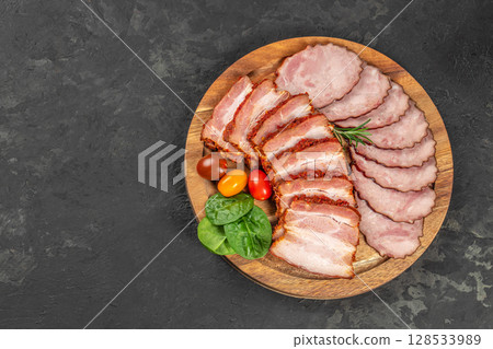 A wooden serving board with a mix of sliced smoked pork and spicy pork belly, garnished with cherry tomatoes, spinach leaves, and rosemary 128533989