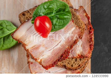 Close-up of an open sandwich with spicy pork belly on rye bread, garnished with a fresh spinach leaf and cherry tomato 128533990