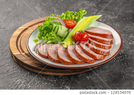 Sliced smoked sausage is elegantly arranged on a plate with fresh lettuce, cucumber ribbons, tomato, and red onion for a vibrant and appetizing presentation 128534070