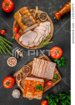 An assortment of sliced deli meats is beautifully arranged on wooden boards with tomatoes, herbs, peppercorns, and salt, creating a rustic and appetizing charcuterie display 128534075