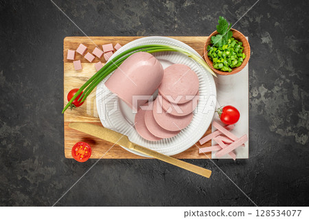 Sliced, diced, and julienned bologna sausage is served on a white plate with fresh green onions, chopped herbs, and cherry tomatoes, creating a vibrant and organized presentation 128534077