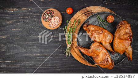 Three perfectly smoked quails are artfully arranged on a dark rustic plate with fresh thyme, coarse salt, and colorful peppercorns, creating a rich and inviting gourmet presentation 128534087