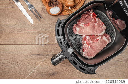 Raw pork chops are placed inside an air fryer basket, surrounded by kitchen tools and seasonings on a wooden countertop, ready for a convenient and healthy cooking process Raw pork chops are placed inside an air fryer basket, surrounded by kitchen tools and seasonings on a wooden countertop, ready for a convenient and healthy cooking process 128534089