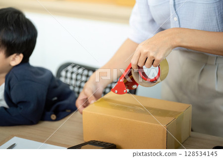 Sell Online: A mother sealing packages for her e-commerce business with her child nearby 128534145