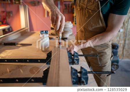 Artisan using screwdriver for clasping clamps to bind wooden beams for large board on workbench 128535029