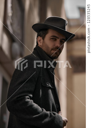 Man in vintage hat and coat standing in sunlit alleyway Man in vintage hat and coat standing in sunlit alleyway 128535245