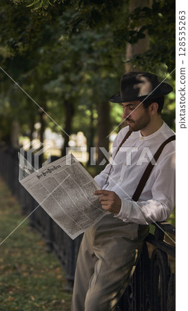 Man reading newspaper in vintage attire leaning against park fence under trees 128535263