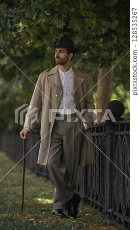 Man in vintage outfit walking by wrought iron fence under trees 128535267
