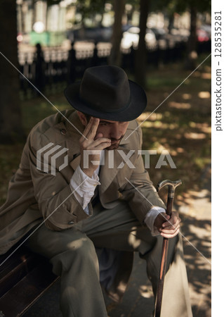 Thoughtful man with hat and cane sitting on park bench in autumn sunlight Thoughtful man with hat and cane sitting on park bench in autumn sunlight 128535281