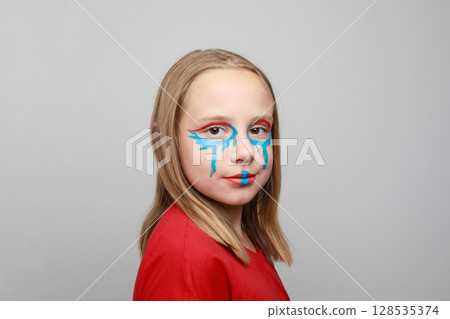 Attractive girl child with vibrant blue and red face paint makeup on her cheeks and forehead. Attractive girl child with vibrant blue and red face paint makeup on her cheeks and forehead. 128535374