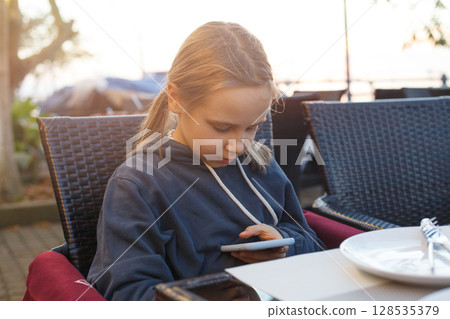 Young girl using smartphone at outdoor cafe during sunset 128535379