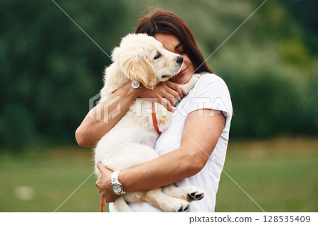 Puppy is in hands. Woman with cute little golden retriever dog is on the green field 128535409