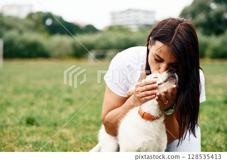 Kissing the animal. Woman with cute little golden retriever dog is on the green field 128535413
