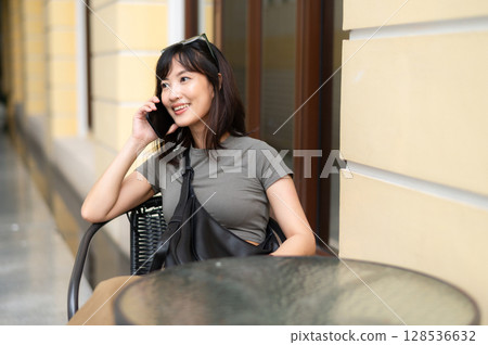 Happy young southeast asian woman blogger in 30s working outside coffee shop room searching internet with smartphone near old architecture building Happy young southeast asian woman blogger in 30s working outside coffee shop room searching internet with smartphone near old architecture building 128536632
