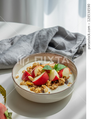Natural greek yogurt with granola and peach in a bowl on a white table near window 128538590