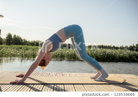 Young woman practicing yoga pose on wooden dock near lake in summer morning light 128538666