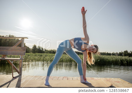 Young woman practicing yoga pose on wooden dock near lake in summer morning light Young woman practicing yoga pose on wooden dock near lake in summer morning light 128538667