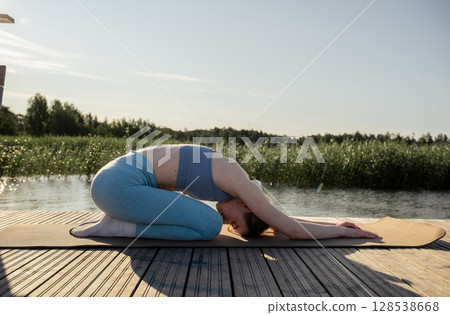 Young woman practicing yoga pose on wooden dock near lake in summer morning light 128538668