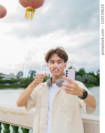 Asian non binary tourist person with headphones relaxing at park near lake using mobile phone Asian non binary tourist person with headphones relaxing at park near lake using mobile phone 128539628
