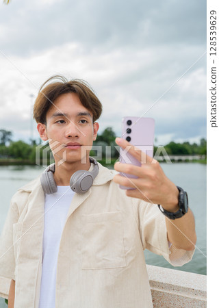 Asian non binary tourist person with headphones relaxing at park near lake using mobile phone Asian non binary tourist person with headphones relaxing at park near lake using mobile phone 128539629