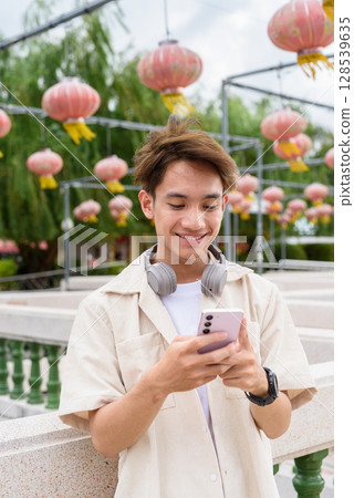 Asian non binary tourist person with headphones relaxing at park near lake using mobile phone 128539635