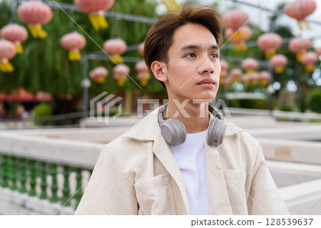 Asian non binary tourist person with headphones relaxing at park near lake 128539637