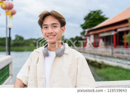 Asian non binary tourist person with headphones relaxing at park near lake 128539643